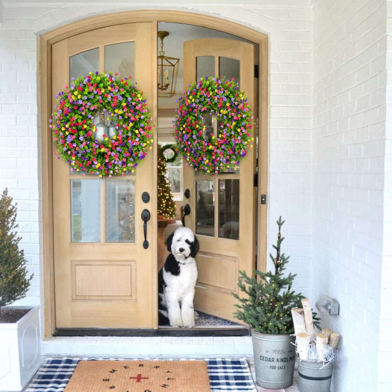 A black and white dog sits in the doorway of a light wood front door decorated with two large, colorful spring flower wreaths. The scene is set on a white brick porch with a blue and white checkered rug, a small potted tree, and a galvanized bucket planter in the foreground.