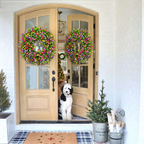 A black and white dog sits in the doorway of a light wood front door decorated with two large, colorful spring flower wreaths. The scene is set on a white brick porch with a blue and white checkered rug, a small potted tree, and a galvanized bucket planter in the foreground.