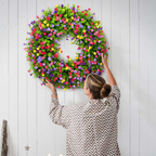 A woman wearing a brown and white chevron shirt hangs a large artificial flower wreath on a white wood-paneled wall.
