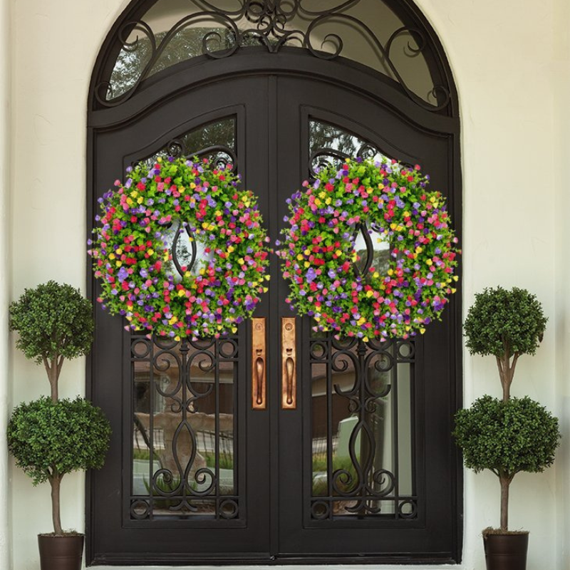 Two large circular floral door wreaths with colorful faux flowers hang on a black wrought-iron front door, flanked by two potted topiary trees against a cream exterior wall.