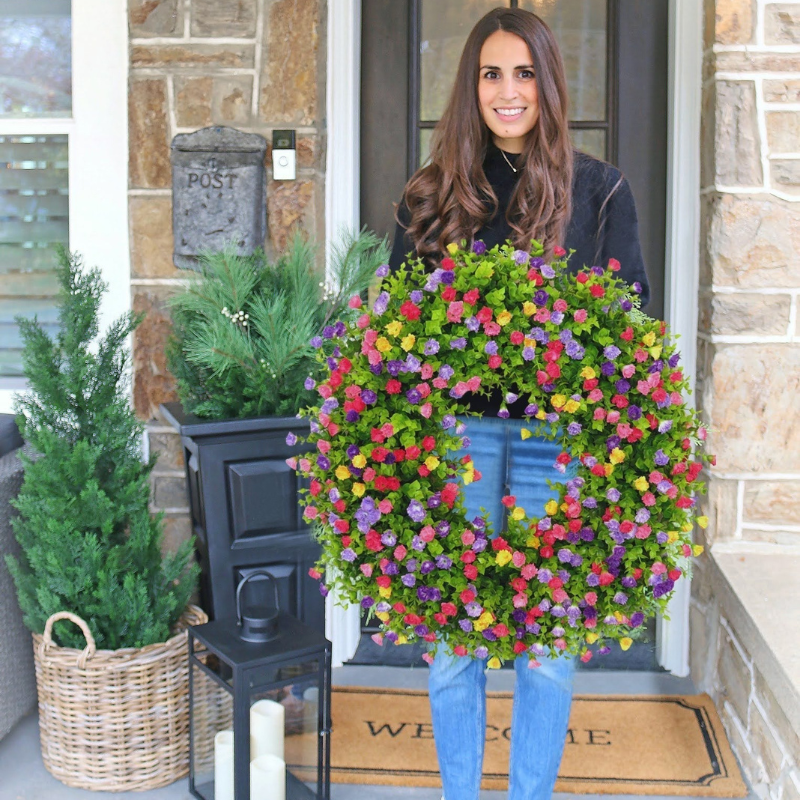 A woman stands on a front porch holding a very large floral wreath made of dense green foliage and many small flowers in pink, purple, yellow, orange, and red. The wreath is wider than the person and covers most of the frame.