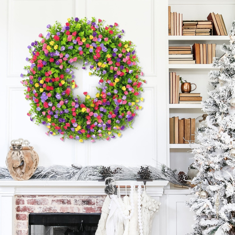 A large artificial floral wreath with green foliage and colorful flowers in pink, purple, yellow, and red, displayed on a white paneled wall above a fireplace mantel in a decorated living room setting.