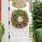 A vibrant, circular artificial flower wreath in shades of pink, purple, yellow, and green, hanging on a white wooden garden gate in a lush garden setting.