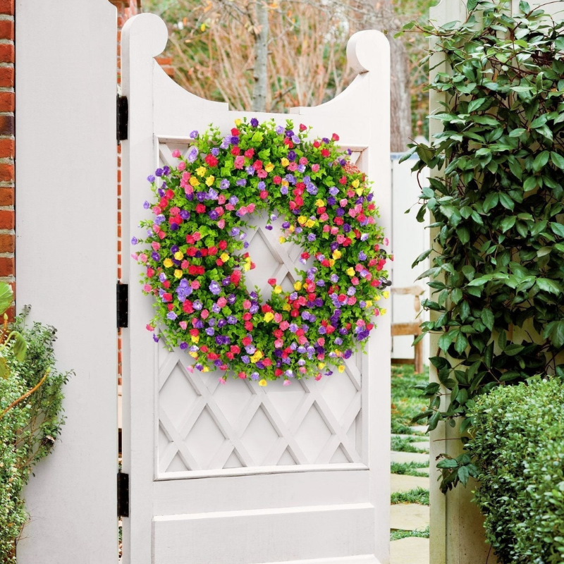 A vibrant, circular artificial flower wreath in shades of pink, purple, yellow, and green, hanging on a white wooden garden gate in a lush garden setting.