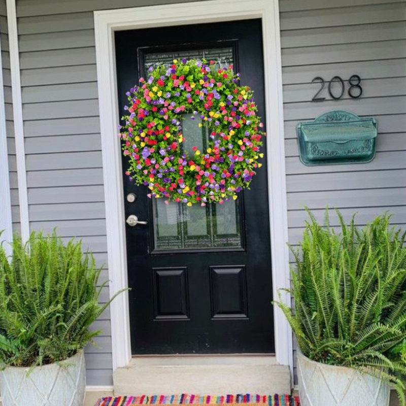 A vibrant, multicolored artificial flower wreath hangs on a black front door of a gray house, flanked by potted green ferns and a green mailbox with the house number 208.
