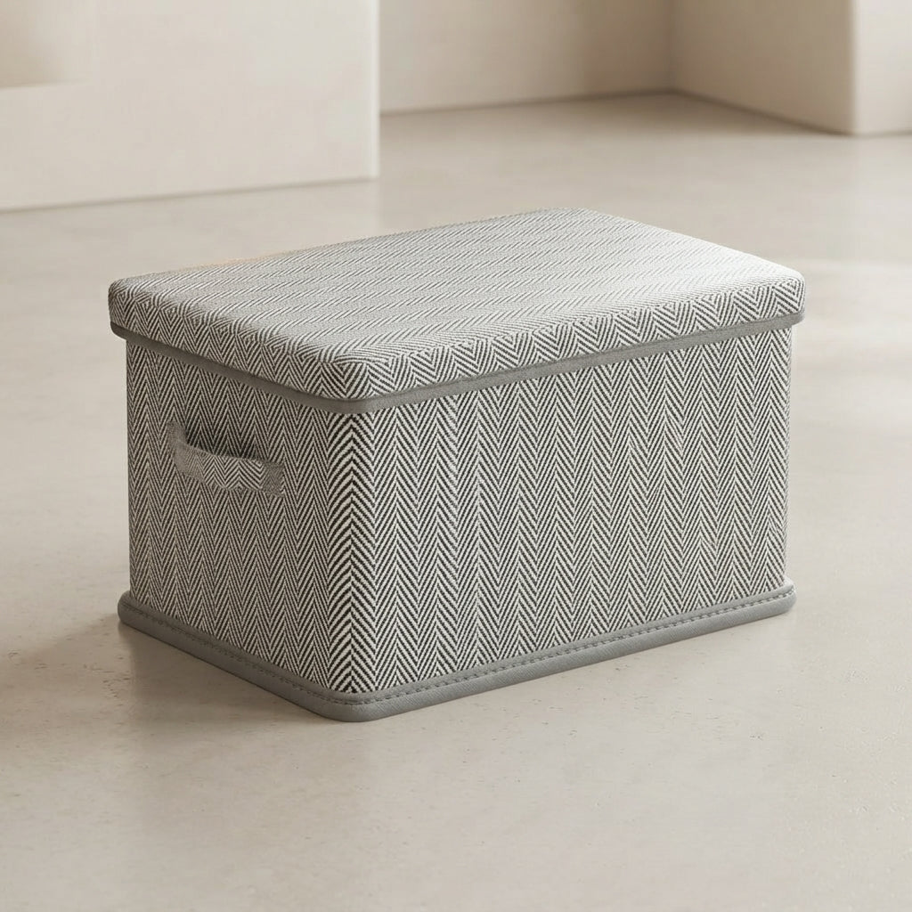 A rectangular storage box with a herringbone-patterned gray and white fabric cover, featuring a small side handle and a gray base, set on a light beige floor.