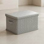 A rectangular storage box with a herringbone-patterned gray and white fabric cover, featuring a small side handle and a gray base, set on a light beige floor.