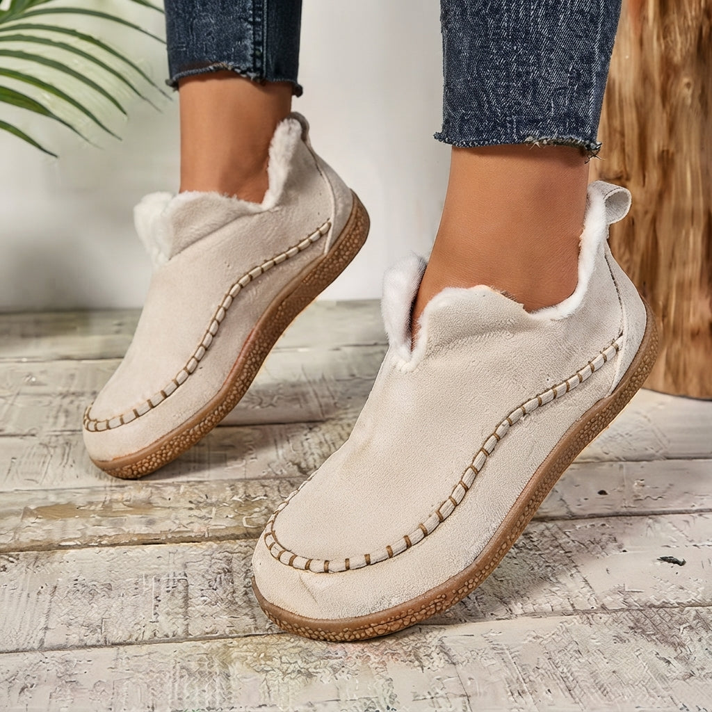A pair of women's slip-on suede moccasin-style slippers in a light beige/cream color with white faux-fur lining and decorative stitching, shown on a person wearing blue jeans on a rustic wooden floor.