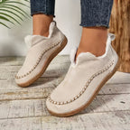 A pair of women's slip-on suede moccasin-style slippers in a light beige/cream color with white faux-fur lining and decorative stitching, shown on a person wearing blue jeans on a rustic wooden floor.