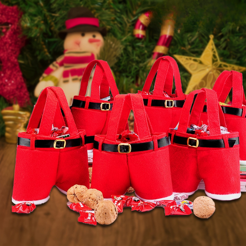 A set of red felt Santa hat-shaped candy baskets with black faux-leather belt details and gold-tone buckle accents, arranged on a wooden surface with wrapped candies and nuts in front. Christmas decorations, including a snowman figure and a star ornament, are blurred in the background.