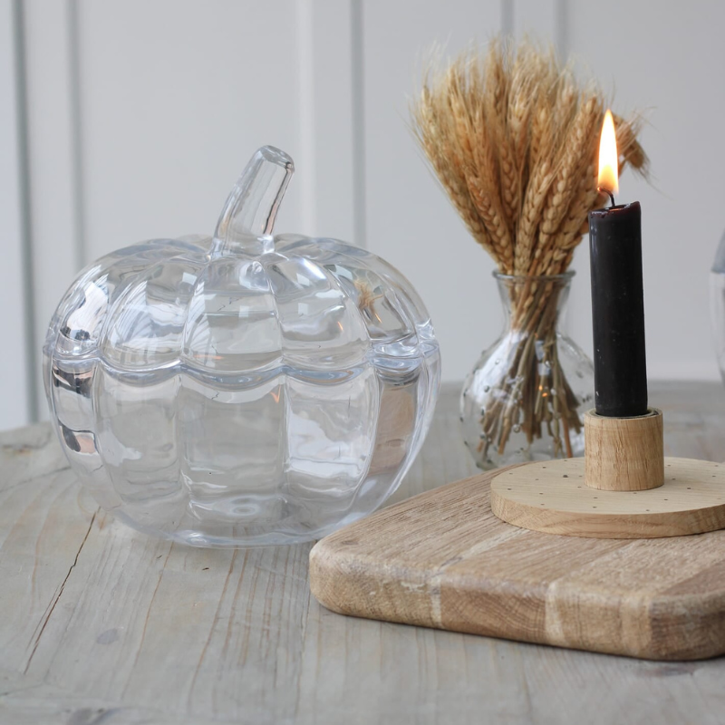 A clear glass pumpkin-shaped container with a ribbed texture and stem, styled on a light wooden table next to a black candle in a wooden holder and dried wheat in a glass vase.