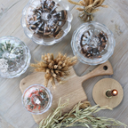 A styled flat-lay of clear glass candle holders and domed glass jars containing dried botanicals, arranged on a light wood cutting board and rustic wood surface with wheat stems and a small candle.