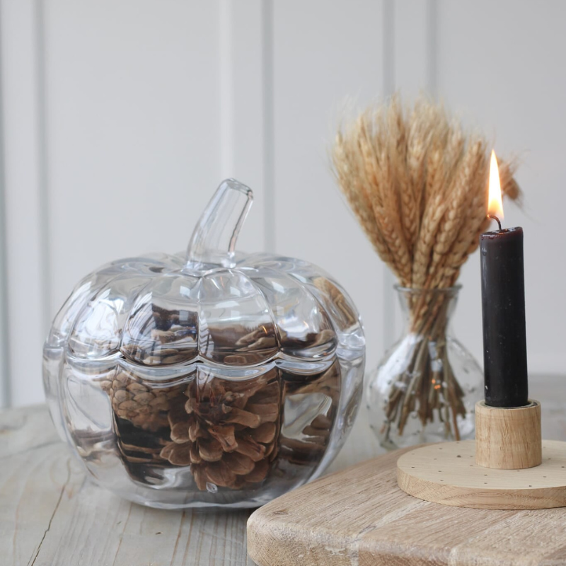 A clear glass pumpkin-shaped container with a stem, filled with pinecones, sits on a wooden surface beside a black candle in a wooden holder and a small glass vase with dried wheat stalks.