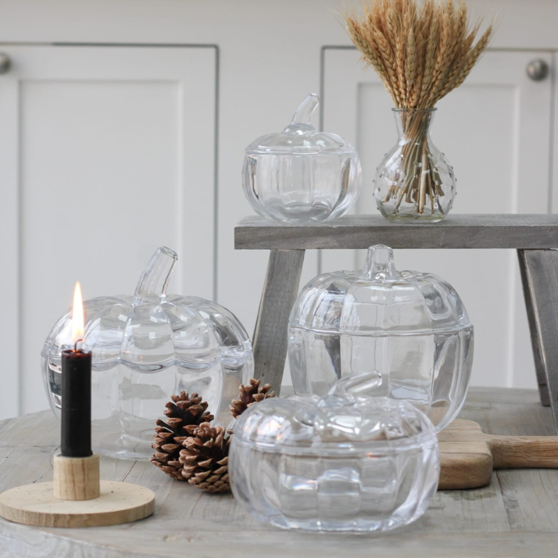 Clear glass pumpkin-shaped lidded containers styled on a rustic wooden table with a small black candle, pinecones, and a small vase of wheat on a gray wooden stool.
