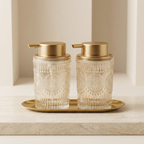 Two glass soap dispensers with gold lids on a matching tray against a neutral background