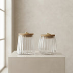 Two ribbed clear glass jars with light wood lids and small gold metal pulls, displayed on a beige pedestal against a neutral beige wall.