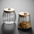 Two ribbed clear glass jars with light wood lids and brass ring handles, one empty and one filled with dried yellow flowers, on a dark gray reflective surface.