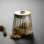 A ribbed clear glass jar with a light wood lid and a small gold ring pull, filled with dried yellow-brown herbs or moss, photographed on a dark gray surface with a few pieces spilled in front.