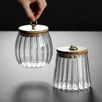 Two ribbed clear glass jars with light wood lids and brass ring pulls, shown on a dark gray surface with a black background. A hand is lifting one jar, and a second jar sits on the table.