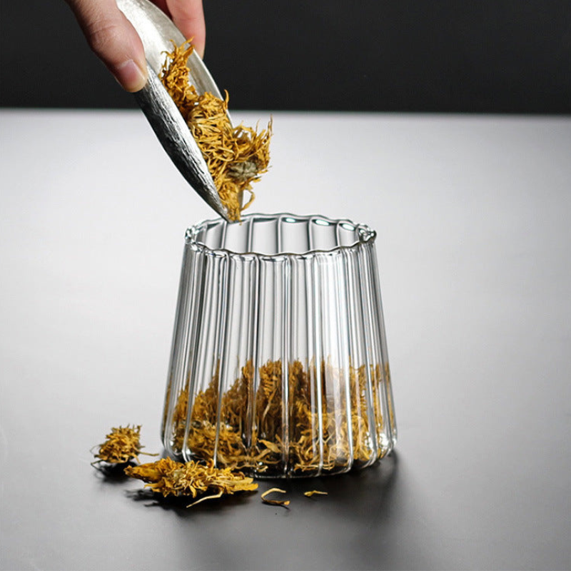 A clear ribbed glass cup on a dark surface, with dried yellow-orange flower buds being poured into it from a metal scoop held by a hand.