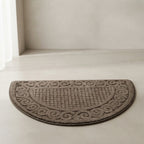 A semi-circular brown bath mat with a raised textured pattern and decorative border, placed on a light beige floor in a minimalist bathroom setting.