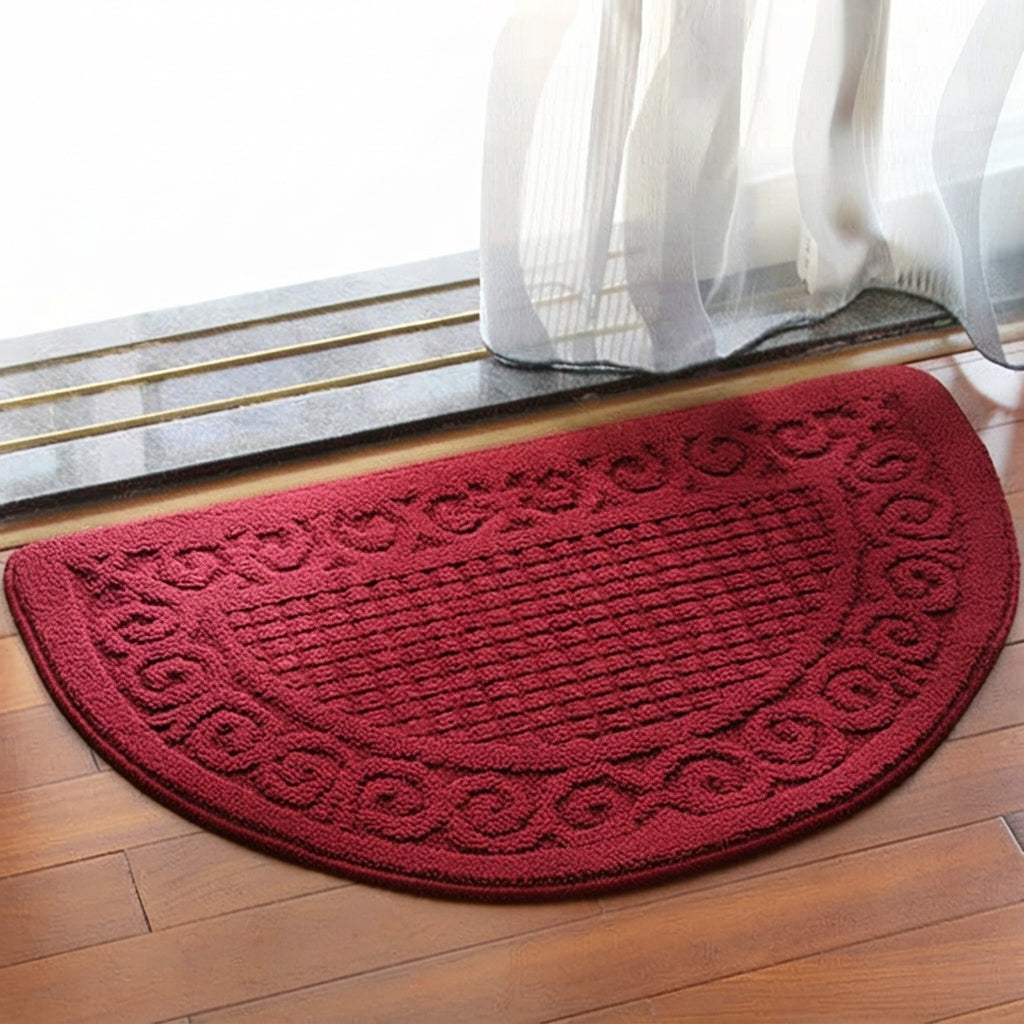 A semi-circular red doormat with a raised decorative border, placed on a wooden floor in front of a glass door with white sheer curtains.