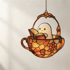 A stained glass hanging ornament shaped like a teacup basket with a smiling ghost reading a book, suspended by a chain against a light wall.
