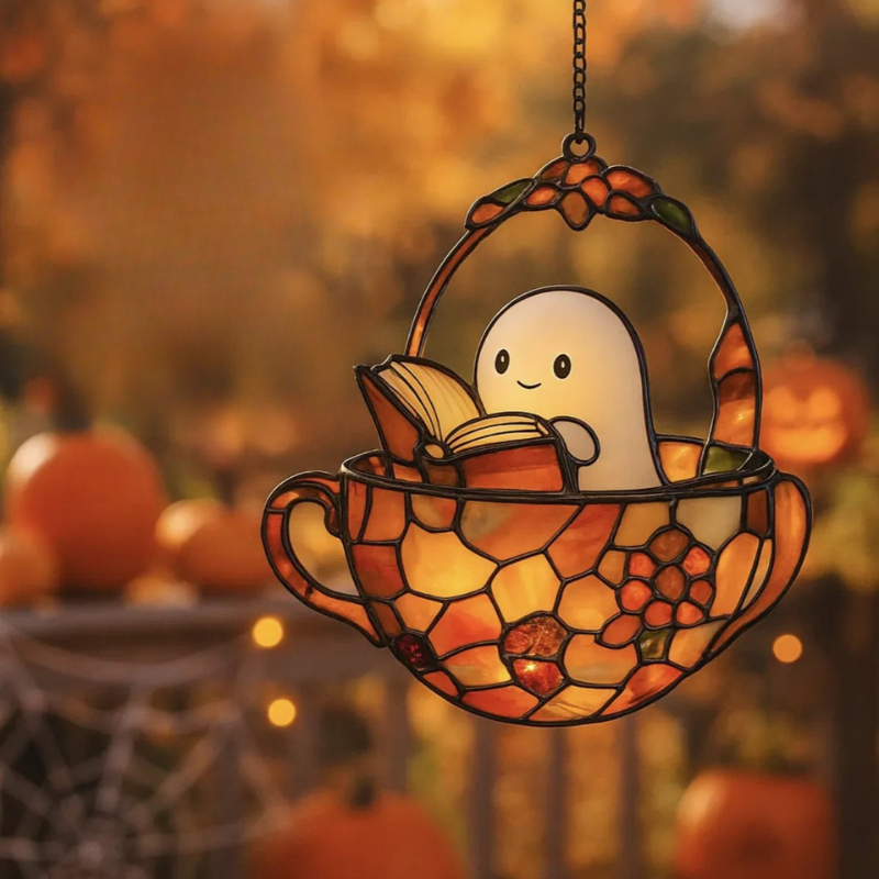 A stained-glass-style hanging ornament shaped like a teacup basket with a small smiling ghost reading a book, shown against a warm autumn outdoor background with pumpkins and bokeh lights.