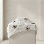 White faux spiderweb draped over a box, decorated with several black plastic spiders, set against a neutral beige background.