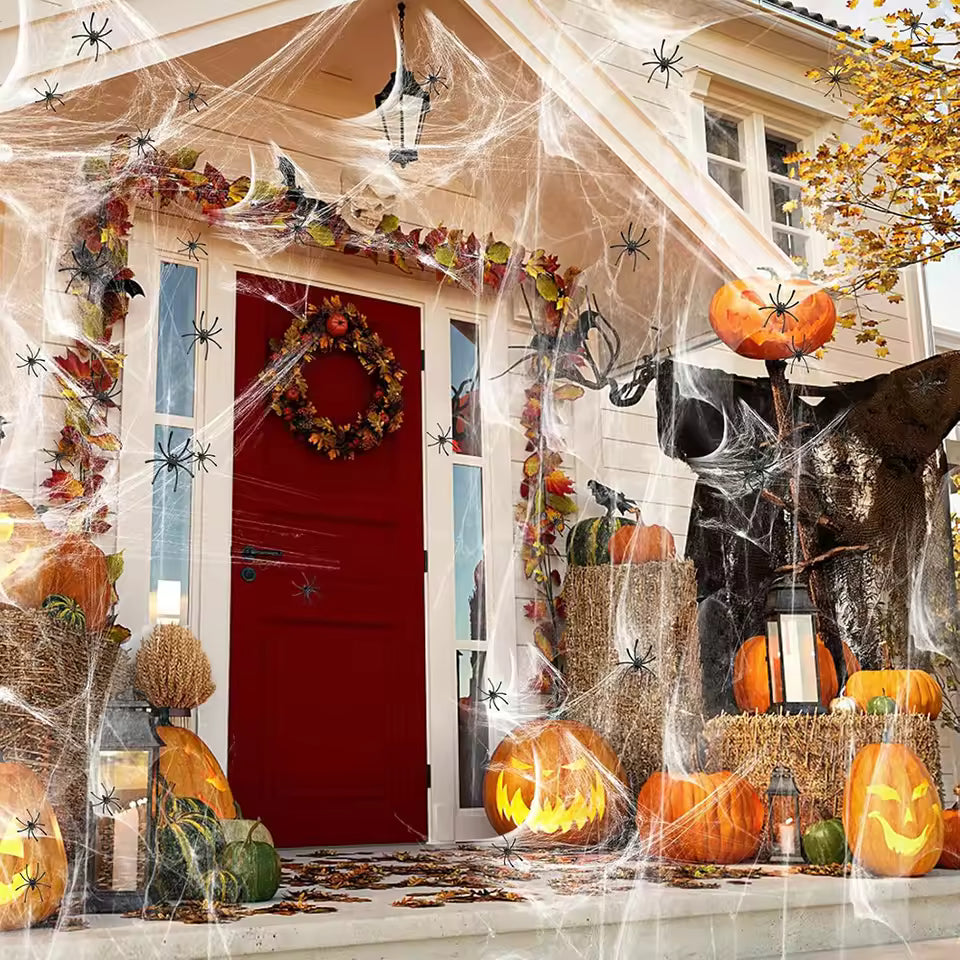A decorated house front porch for Halloween, with a red front door, many orange pumpkins including carved jack-o'-lanterns, cobwebs draped across the porch and roof, spider decorations, fall leaves, hay bales, and a large black bat hanging on the right.