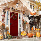 A decorated house front porch for Halloween, with a red front door, many orange pumpkins including carved jack-o'-lanterns, cobwebs draped across the porch and roof, spider decorations, fall leaves, hay bales, and a large black bat hanging on the right.