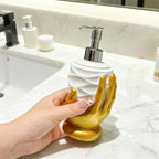 A hand holds a white and gold soap dispenser shaped like a sculpted hand with a chrome pump, set on a white marble bathroom counter.