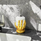 A bathroom soap dispenser with a white cylindrical body and a sculpted gold hand base, topped with a silver pump, sitting on a dark countertop near a sink.