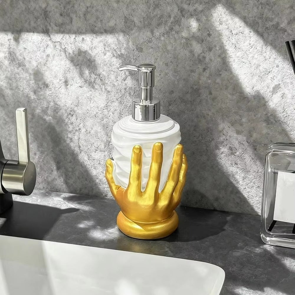 A bathroom soap dispenser with a white cylindrical body and a sculpted gold hand base, topped with a silver pump, sitting on a dark countertop near a sink.