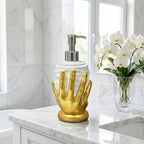 A bathroom soap dispenser with a white cylindrical body and a sculpted metallic gold hand-shaped base, topped with a silver pump. It sits on a white marble countertop next to a vase of white orchids.