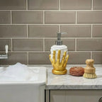 A bathroom countertop scene with a gold hand-shaped soap dispenser holding a white bottle and silver pump, next to a wooden scrubber and sponge on a marble counter, with a tiled backsplash behind.