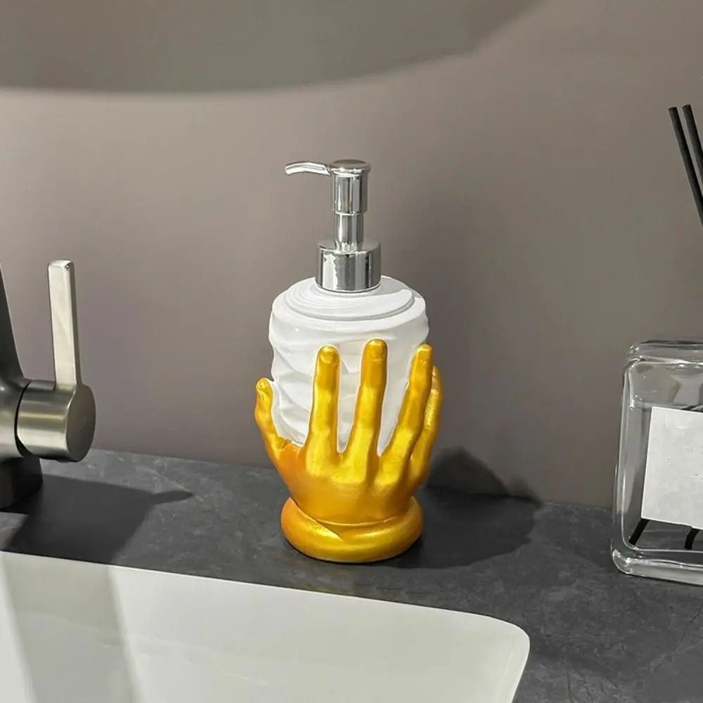 A decorative soap dispenser with a white ribbed cylindrical body and a metallic gold hand-shaped base, shown on a bathroom counter beside a faucet and glass container.