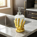 A decorative soap dispenser with a white ribbed cylindrical body and a metallic gold hand-shaped base, placed on a white marble kitchen countertop next to a stainless steel sink.