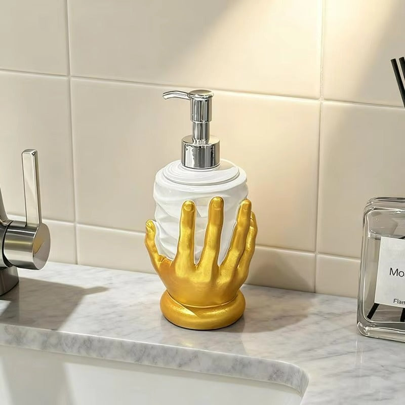 A decorative soap dispenser shaped like a glossy gold hand holding a white cylindrical bottle, set on a marble bathroom counter with a tiled wall behind it.
