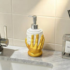A decorative soap dispenser shaped like a glossy gold hand holding a white cylindrical bottle, set on a marble bathroom counter with a tiled wall behind it.