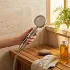 Handheld shower head held by a hand in a bathroom setting with a wooden bench, towel, and bottle of oil.