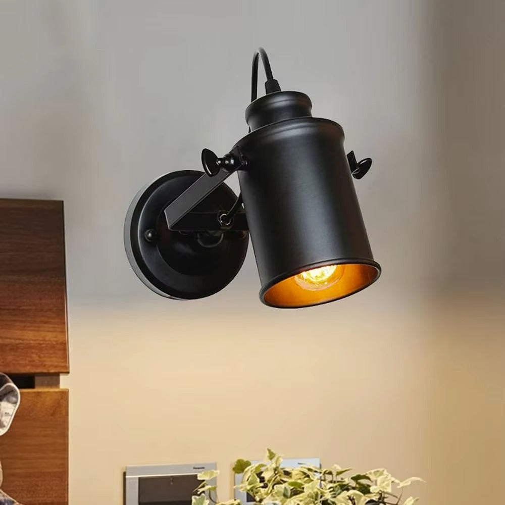 A black industrial-style wall sconce with a cylindrical shade and adjustable arm, shown lit against a beige wall with a wooden cabinet and plant visible in the room.