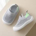A pair of light gray knit slip-on baby shoes with white soles and a small neon green accent on the heel, displayed on a beige surface.