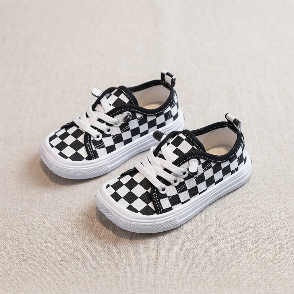 A pair of toddler canvas sneakers with a black and white checkerboard pattern, white laces, and white rubber soles, displayed on a textured beige fabric background.