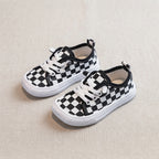 A pair of toddler canvas sneakers with a black and white checkerboard pattern, white laces, and white rubber soles, displayed on a textured beige fabric background.