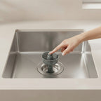 A stainless steel kitchen sink with a gray drain filter being held by a person's hand, shown in a clean, modern kitchen setting.