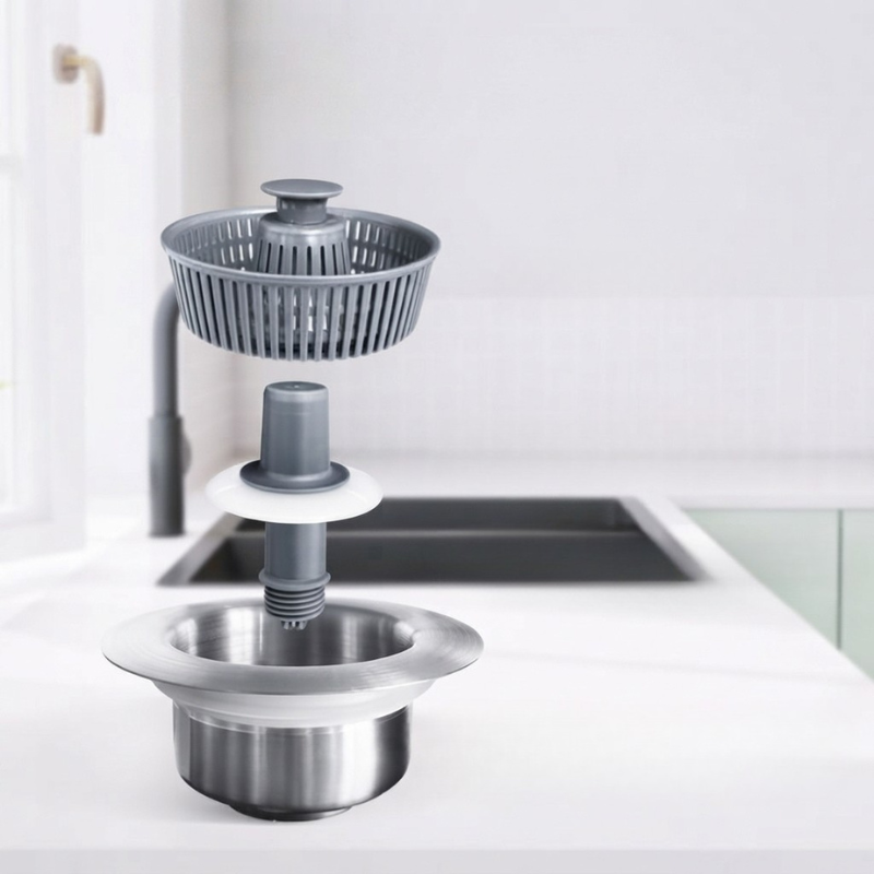 A sink drain assembly with a gray strainer basket, gray central plug, white spacer ring, and stainless steel basin, shown installed on a white kitchen sink countertop with a blurred kitchen background.