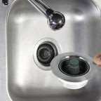 A close-up of a stainless steel kitchen sink with a chrome faucet, showing a hand holding a round drain cover above the drain opening.