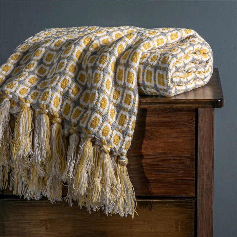 Yellow and gray patterned blanket with tassels draped over a wooden bench against a gray wall.