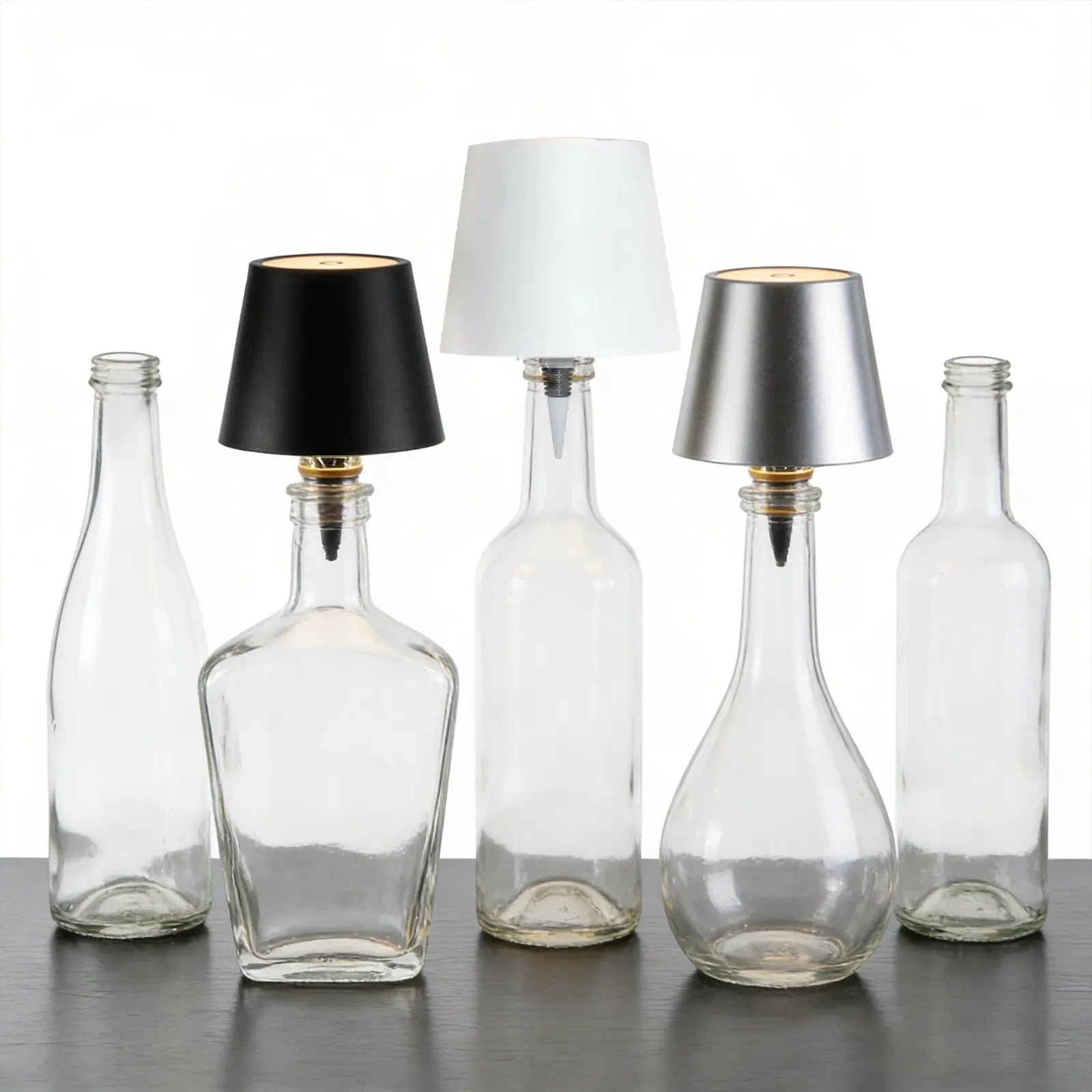 Clear glass bottles with black, white, and silver lampshades on a reflective surface.